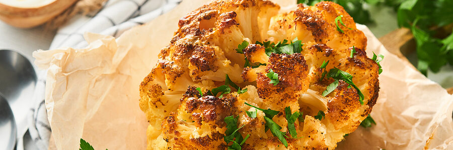 A whole roasted cauliflower garnished with chopped parsley, served on a wooden board with parchment paper.